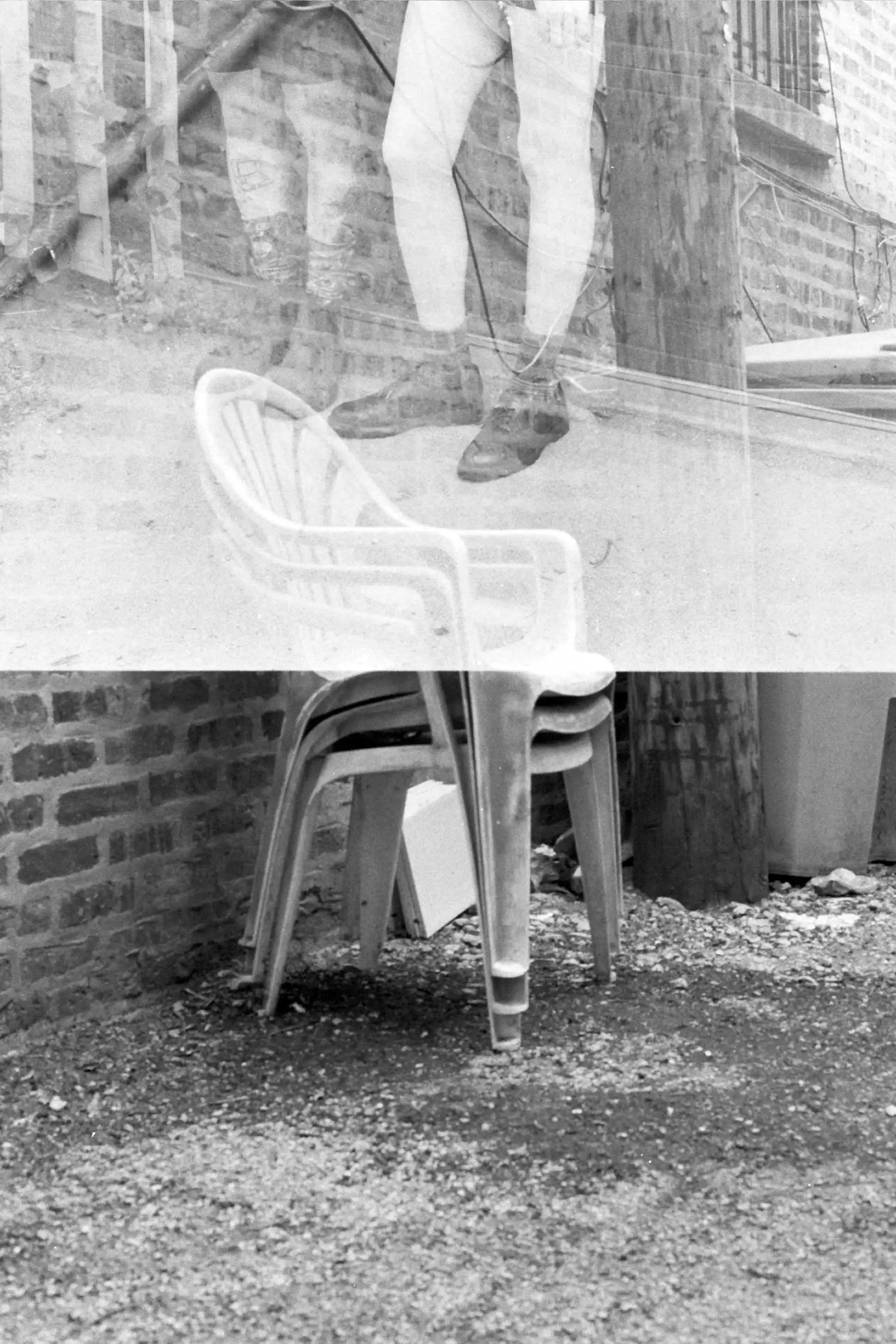 An accidental double exposure. The first exposure covers only the top half of the frame and shows legs of two people standing against a wall outdoors. The second exposure covers the whole frame and shows a stack of plastic chairs against a brick wall in an alley next to a telephone pole. This photo is in black and white.