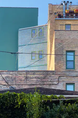 a large multi-story building with a facade made of glass bricks sits nestled between a painted brick seafoam green building and an unpainted yellow brick building. Electrical wires crisscross in the foreground
