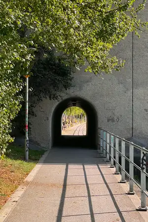 a pedestrian tunnel with an arched roof through a large bridge that spans a river on the right. There's a fence on the right between the sidewalk and the river.