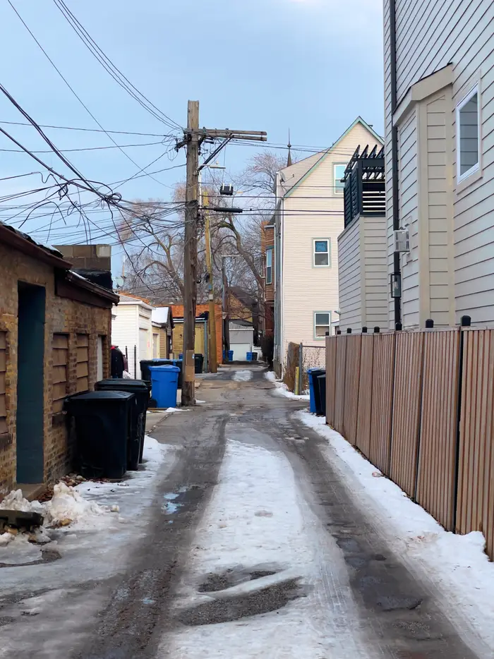 a snow-covered Chicago alley with slushy tire tracks. The alley is lined on one side by a wooden fence