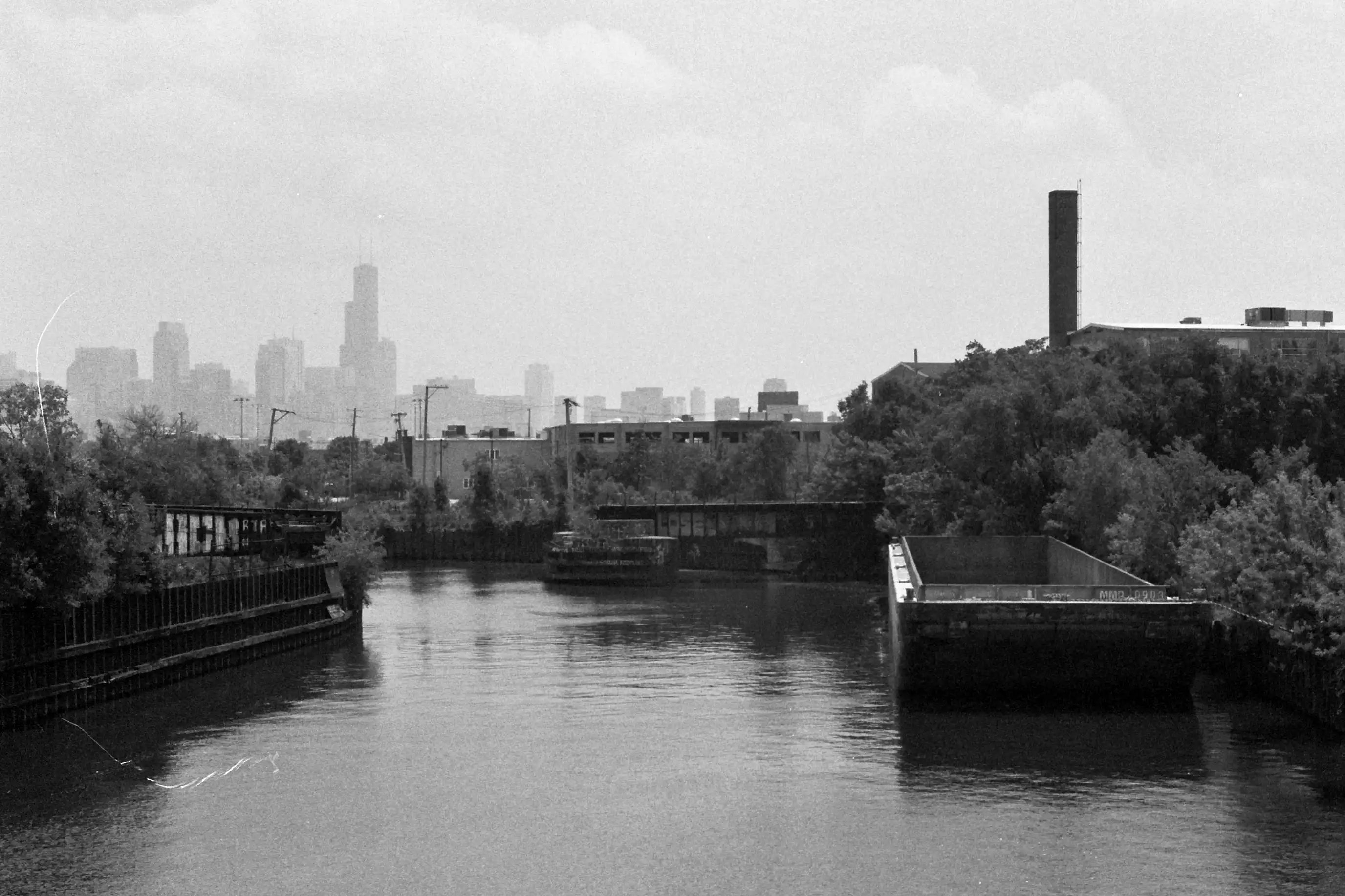 A view of the north branch of the Chicago river looking towards the Chicago skyline. An empty barge floats on the right side of the frame. This photo is in black and white.