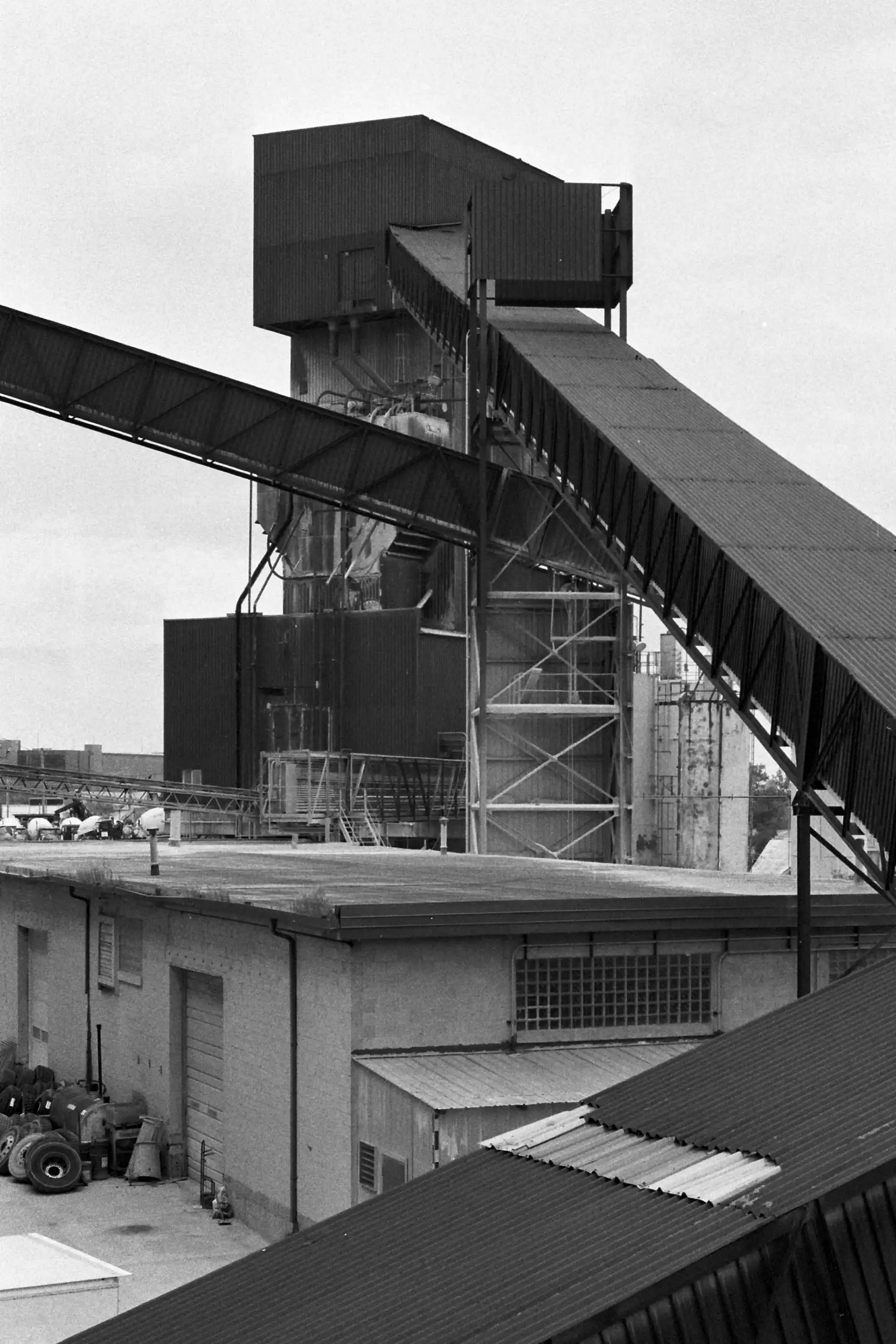 Industrial structures, dark conveyor belts, a central tower. The conveyor belt on the right extends down from the tower to the right side of the frame. Another cuts across the frame from just below the top of the tower and the other conveyor belt  to the left of the frame. A low building with a garage door sits below the tower. This photo is in black and white.