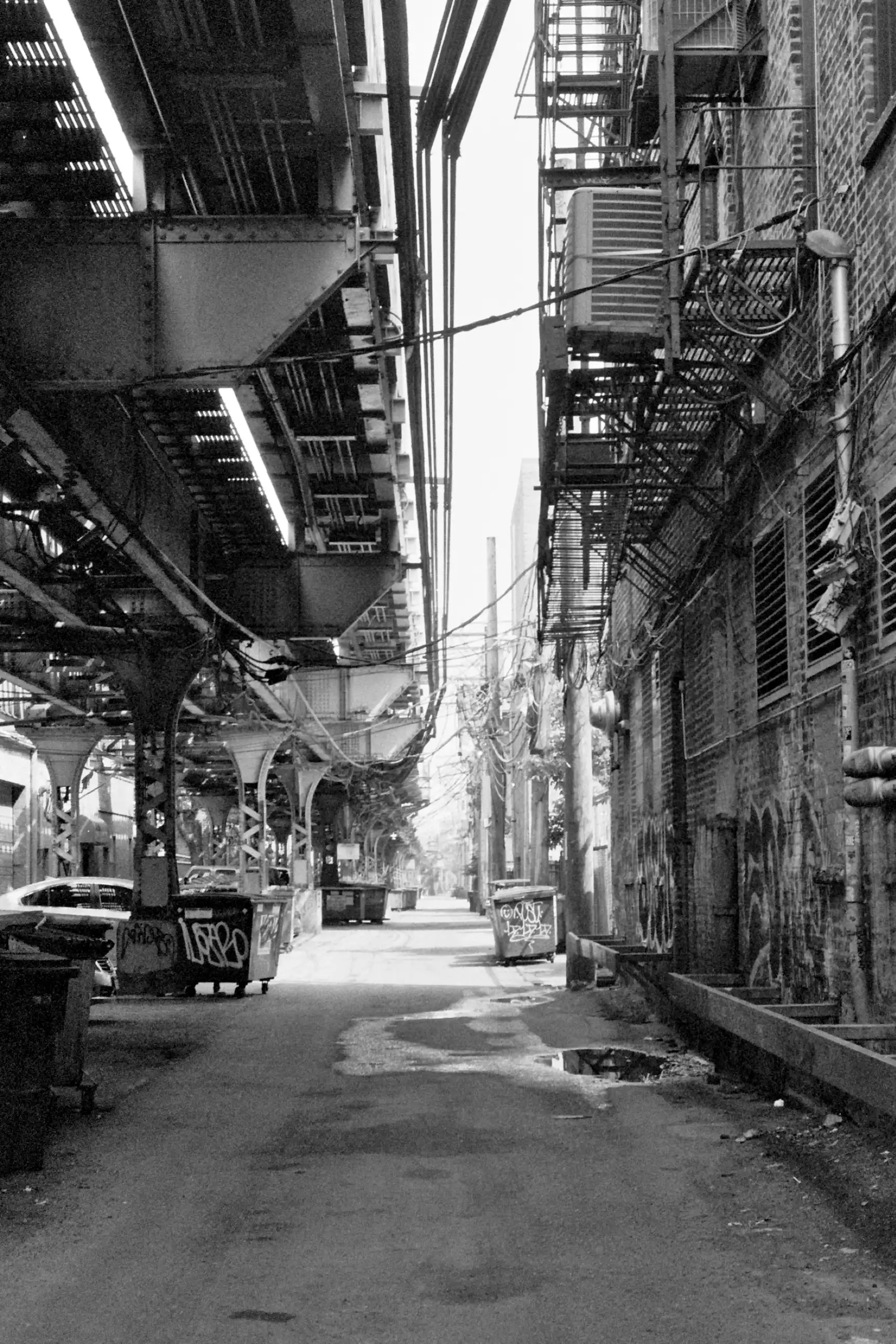 On the left side of the frame is an elevated Chicago transitway. On the right side is the back of a building with fire escapes on its walls. The center of the frame, in the distance is illuminated by sunlight. The foreground is in shadow. Dumpsters and a car are visible in the alley. This photo is in black and white.