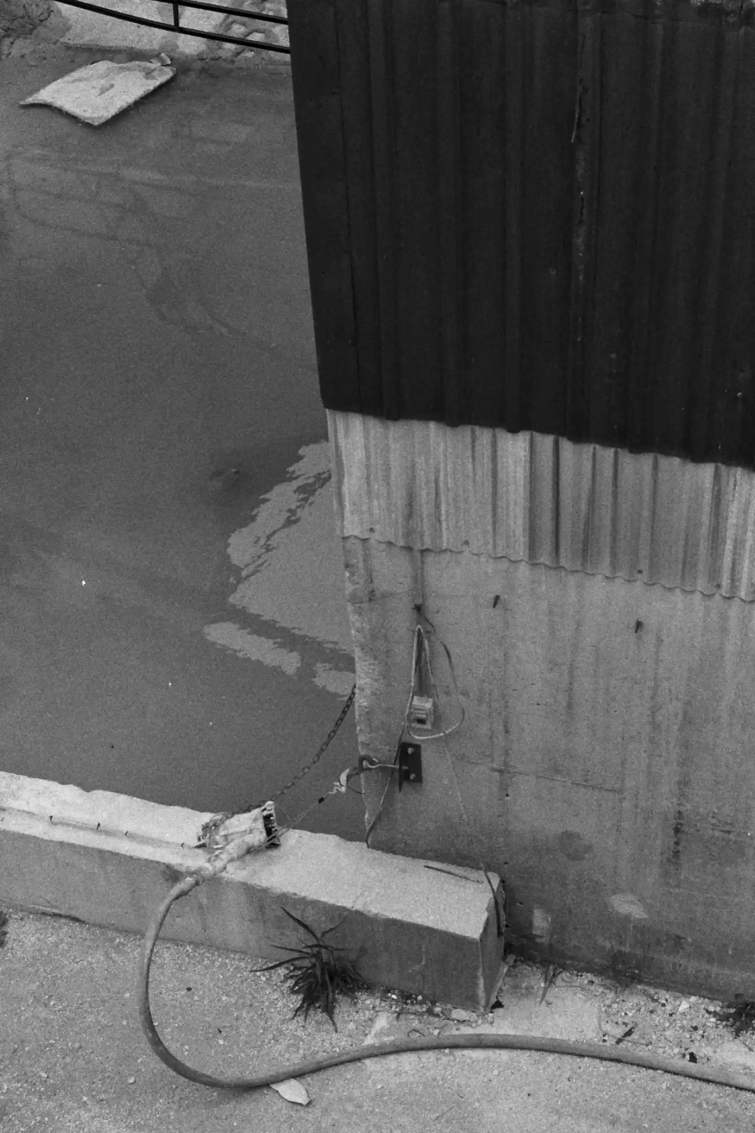 The corner of an industrial building where it meets the ground. On the ground, in front of the building is an unplugged hose of some type. The left of the building is standing water, possibly flooded. The top half of the visible portion of the building is dark sheet metal, the bottom half is light concrete. This photo is in black and white.