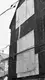 Building with dark burned wooden cladding with a few large panels of light plywood attached to the side. Electrical wires run diagonally across the frame in front of the building. This photo is in black and white.