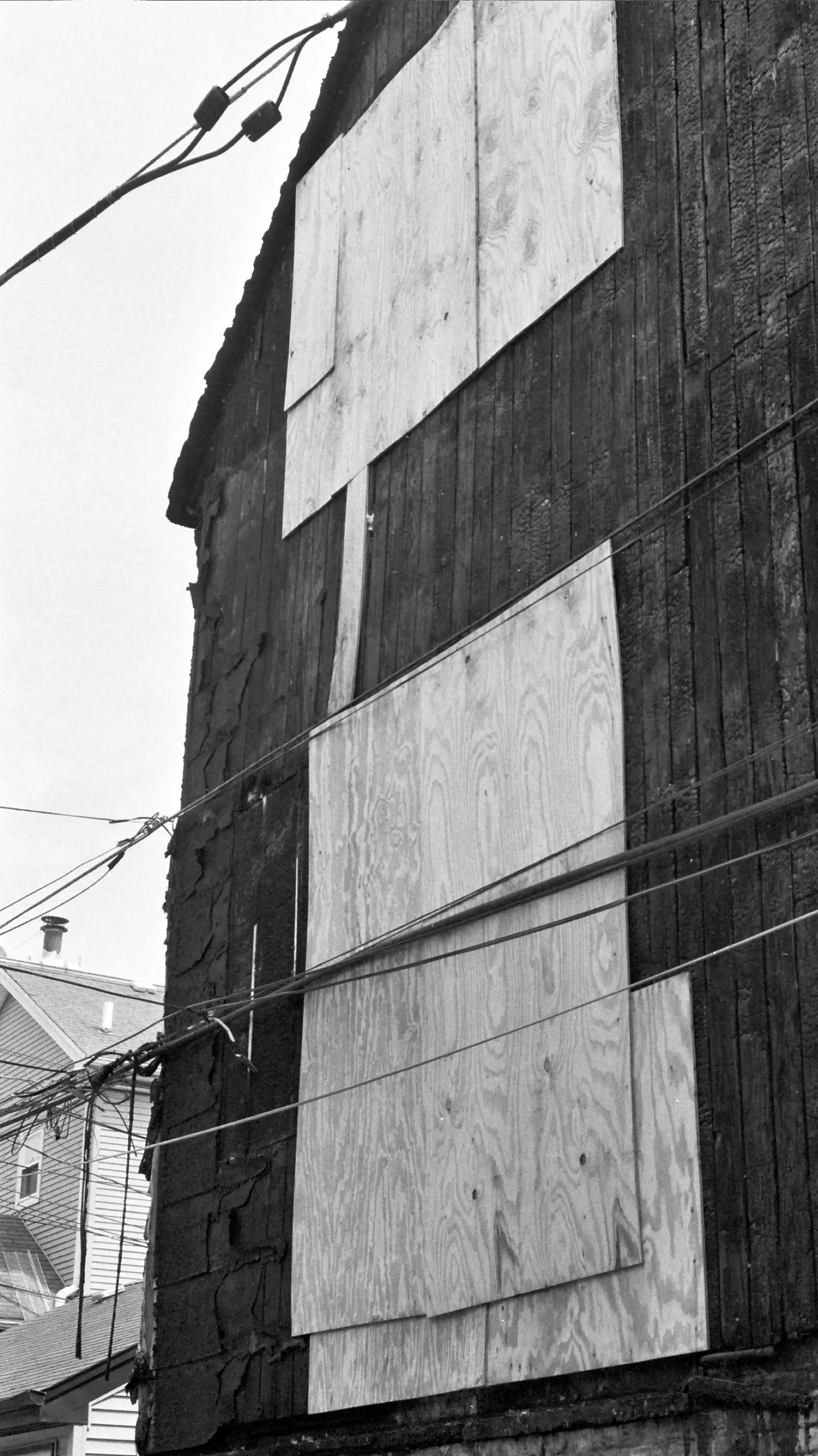 Building with dark burned wooden cladding with a few large panels of light plywood attached to the side. Electrical wires run diagonally across the frame in front of the building. This photo is in black and white.