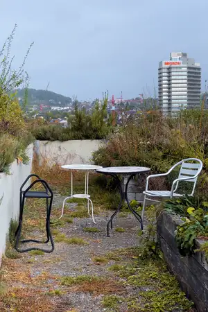 a chair and three small tables are situated on a rooftop in Zurich on an overcast day. The rooftop is bordered by box planters which are overgrown with grasses and other plants.